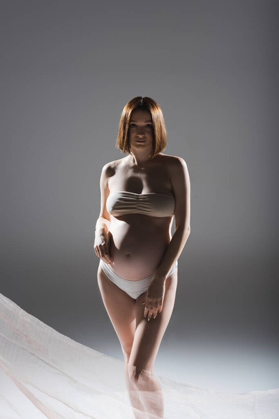 Schwangere in Unterwäsche posiert in Stoffnähe auf grauem Hintergrund - Foto, Bild