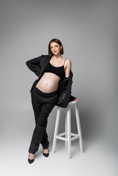 Terhes nő öltönyben és sarkú pózol közelében szék szürke háttér - Fotó, kép