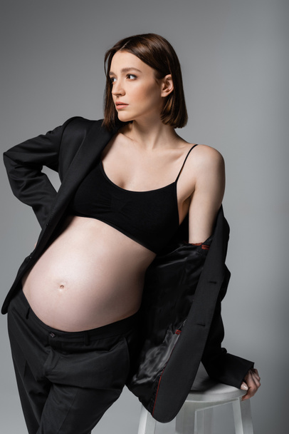 Stylish pregnant woman in suit looking away near chair isolated on grey  - Fotografie, Obrázek
