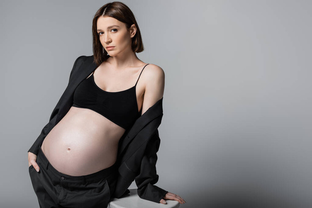 Pregnant woman in top and suit standing near chair isolated on grey  - Fotó, kép
