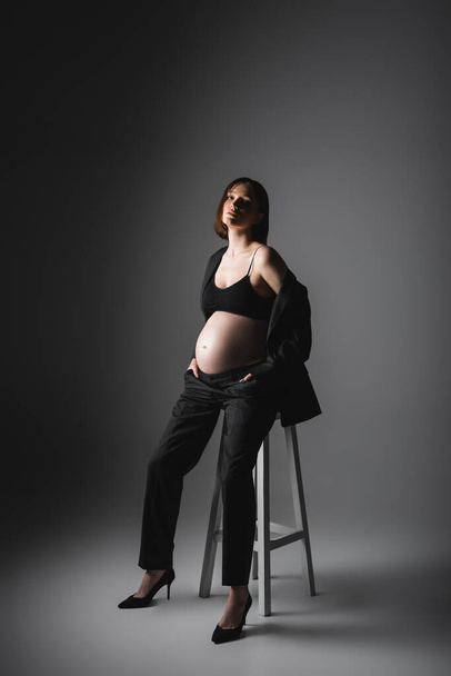 Stijlvolle zwangere vrouw in hoge hakken en jas zitten op stoel op donkergrijze achtergrond  - Foto, afbeelding