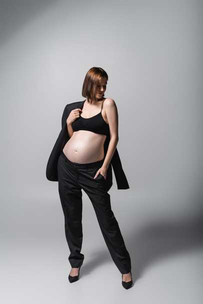 Donna incinta alla moda in tacchi che tiene blazer su sfondo grigio - Foto, immagini