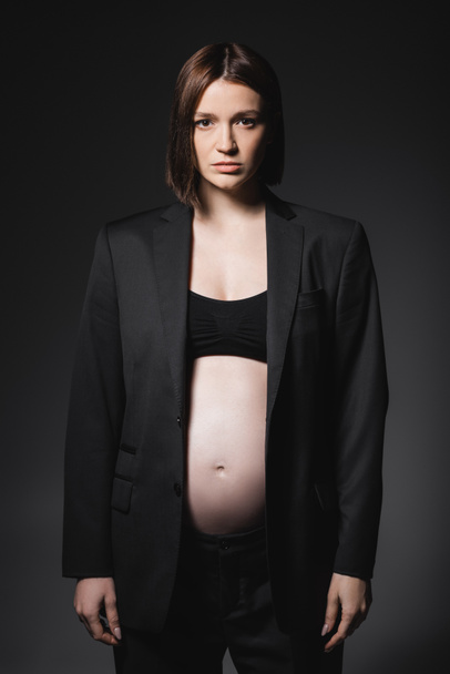 Mujer embarazada en la parte superior y chaqueta mirando a la cámara aislada en gris oscuro  - Foto, imagen