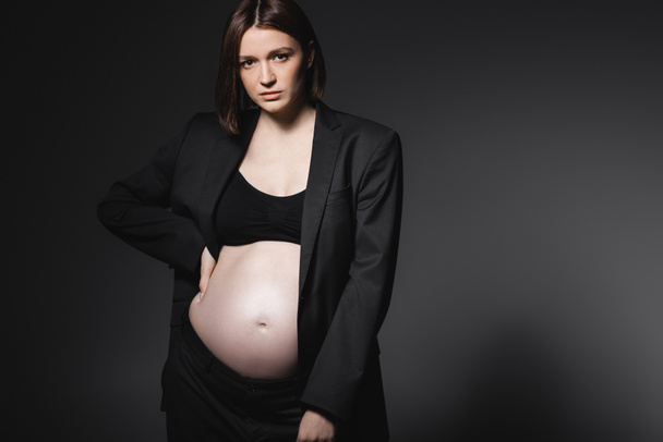 Fashionable pregnant woman in black blazer holding hand on hip isolated on dark grey  - Foto, Bild