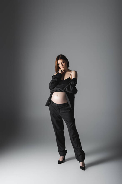 Full length of pregnant woman in heels and suit standing on grey background  - Φωτογραφία, εικόνα