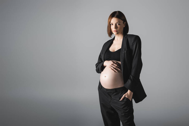 Mulher grávida em blazer e barriga de toque superior isolado em cinza  - Foto, Imagem