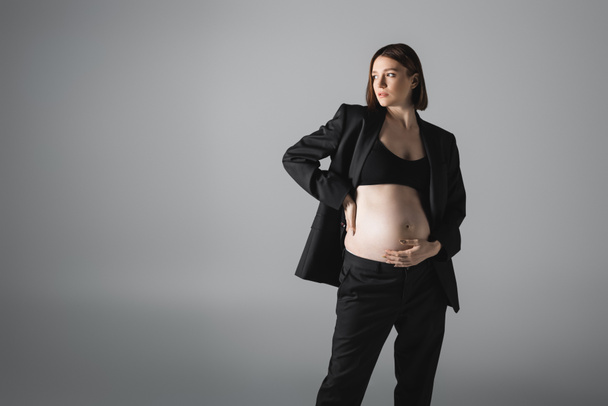 Stylish pregnant woman in top and jacket touching belly isolated on grey  - Фото, изображение