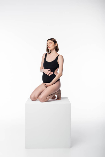 Pregnant woman with closed eyes posing in swimsuit on cube on white background - Φωτογραφία, εικόνα