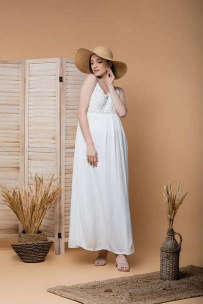 Young pregnant woman in straw hat and dress standing near folding screen and spikelets on beige  - Photo, Image