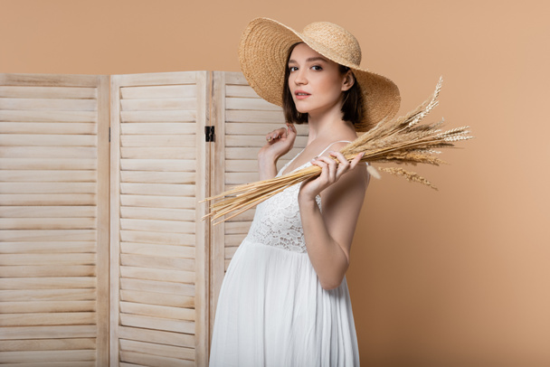 Pregnant woman in sun hat holding spikelets and looking at camera isolated on beige  - Photo, Image