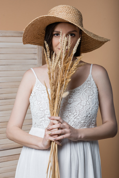 Pregnant woman in sun hat holding spikelets near face isolated on beige  - Foto, Imagen
