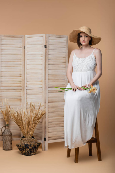 Pregnant woman in sun hat holding flowers while sitting near folding screen on beige  - Foto, immagini