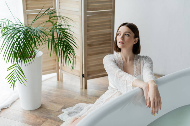 Mujer joven en bata mirando hacia otro lado cerca de la bañera y planta en el baño  - Foto, Imagen