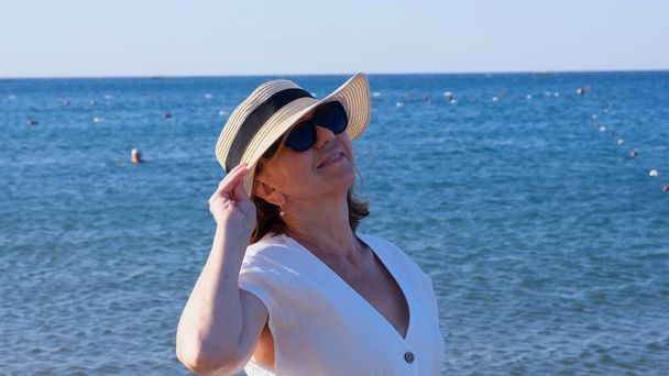 Portré egy elégedett 50 éves nőről, aki szalmakalapot és napszemüveget visel, és élvezi a napot a kék tenger ellen. Nyár, nyaralás, vakáció, aktív nyugdíjasok - Fotó, kép