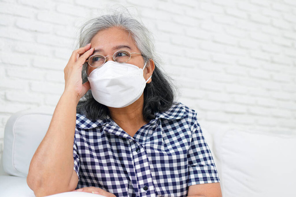 An elderly Asian woman wearing a mask is sitting on the sofa in her home. She was worried about her health problems and the coronavirus. There is no one to take care of. Elderly health care. - Photo, Image