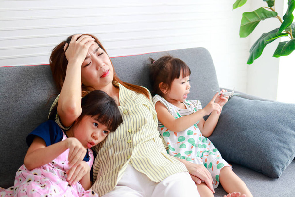 アジアのシングルマザーは2人のいたずら好きな小さな子供を育てる頭痛。三人は家の中のソファに座った。家族の概念。現代の子育て、シングルマザー、幸せな子供を育てる方法. - 写真・画像