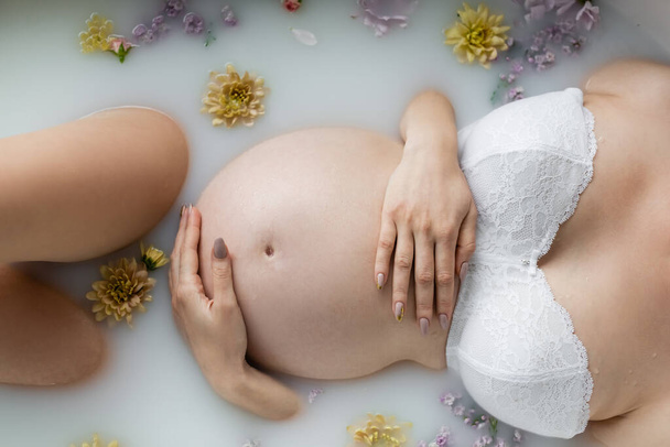 Cropped view of pregnant woman in bra lying in milk bath with plants at home  - Photo, image