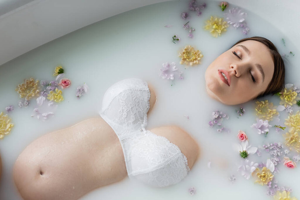 Top view of pregnant woman with closed eyes relaxing in milk bath with flowers  - Фото, изображение