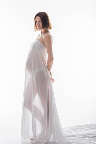 Full length of barefoot pregnant woman posing in fabric on white background  - Foto, immagini
