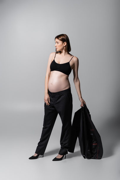 Side view of pregnant woman in top holding jacket on grey background - Fotografie, Obrázek