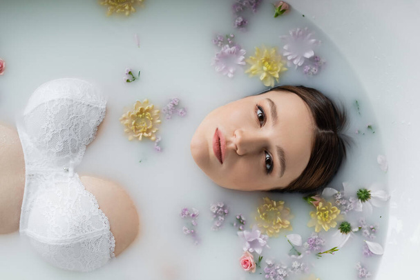 Top view of pretty woman looking at camera while taking milk bath with flowers  - Foto, Imagen