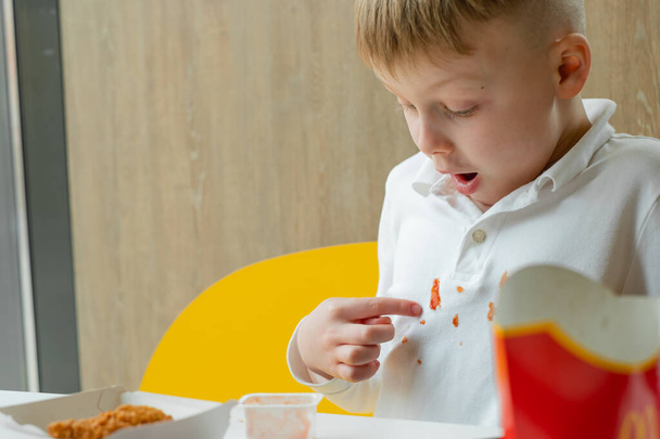 The boy points a finger at the ketchup stain on his clothes at the table in a fast food restaurant. Kids eat unhealthy fat food. The concept of cleaning stains on clothes. - Φωτογραφία, εικόνα