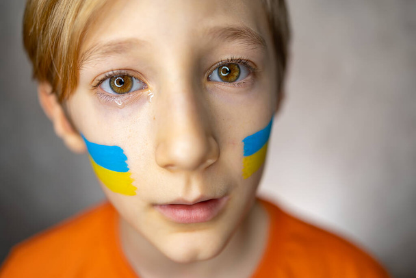 ένα παιδί με θλιμμένα μάτια στο φόντο του πολέμου, ένα αγόρι με μια ζωγραφισμένη σημαία της Ουκρανίας στα μάγουλά του κοιτάζει την κάμερα με δάκρυα - Φωτογραφία, εικόνα
