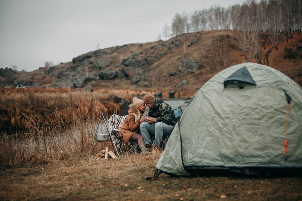 A couple in love in autumn on a camping near the river, sitting on chairs, looking at each other with mugs of tea in their hands. - Valokuva, kuva