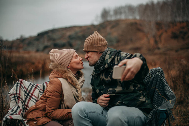 A couple in love in the fall on a camping near the river, sit on chairs and take a selfie on a smartphone - Photo, Image