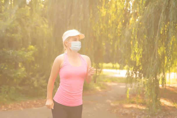Pandemic coronavirus COVID-19 A woman in a meadow in a sports outfit wearing a protective mask in case of the spread of SARS-CoV-2 disease virus. Girl with a protective mask on her face. - Фото, изображение