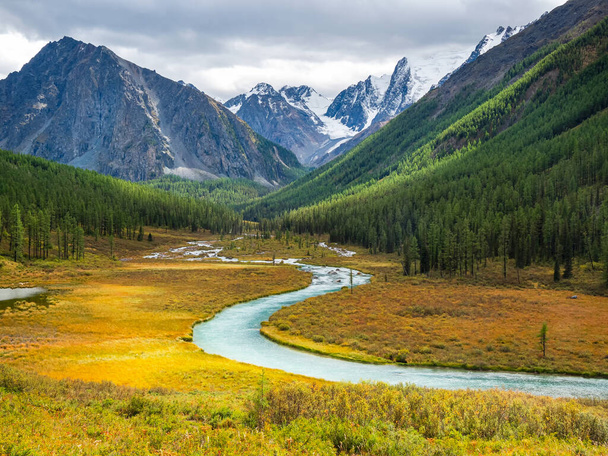 Bright sunny autumn landscape with winding river and  sunlit gold valley with green fir trees on mountainside under cloudy sky. Awesome alpine scenery with beautiful mountains in golden sunshine.  - Photo, image