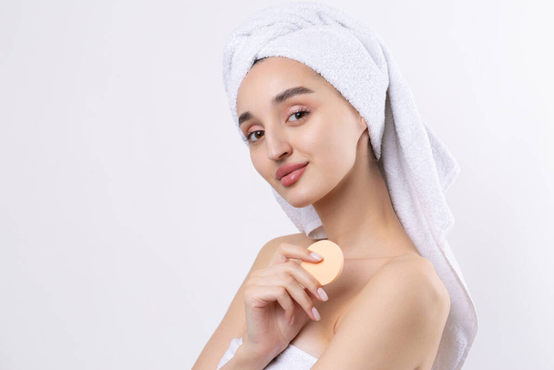 Beautiful girl with thick eyebrows and perfect skin at white background, towel on head, beauty photo. Holds a cosmetic sponge. - Photo, Image