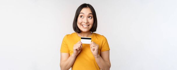 Retrato de hermosa chica coreana sosteniendo tarjeta de crédito, recomendando servicio bancario, de pie en camiseta amarilla sobre fondo blanco - Foto, imagen