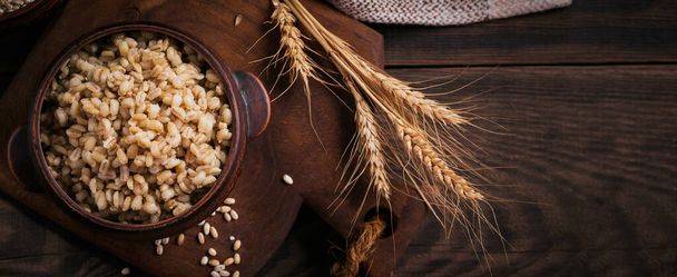 Bowl of cooked peeled barley grains porridge with ears of wheat on dark wooden background. Cooking Healthy and diet food concept. - Foto, immagini