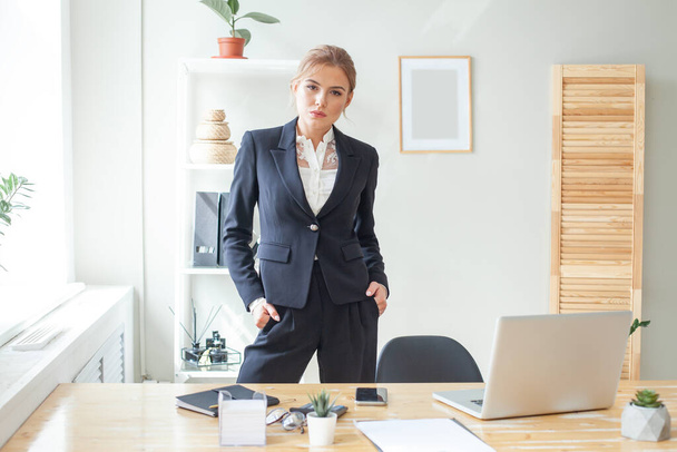 Уверенный эксперт по бизнесу. Привлекательная деловая женщина в умной черной одежде смотрит в камеру, стоя возле своего стола в креативном офисе - Фото, изображение