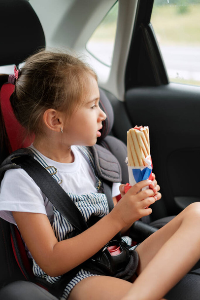 A child in a car seat, fastened with seat belts, holds a French bun with a sausage in his hands. The girl got hungry on the trip and eats a hot dog. Takeaway food. Food drive. - Photo, Image