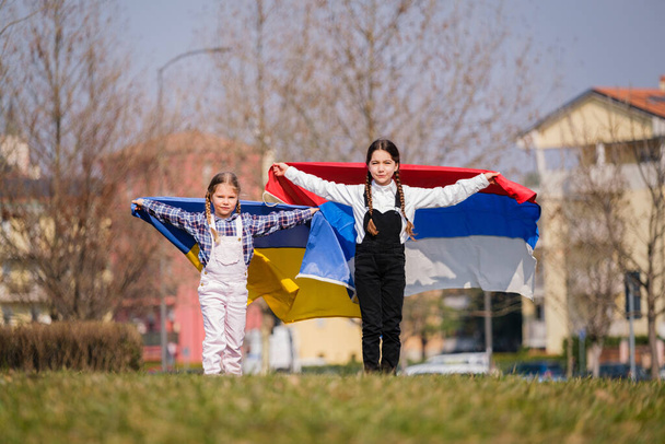 Όμορφα κοριτσάκια με Ουκρανικές και Ρωσικές σημαίες τρέχουν χαρούμενα στο πάρκο. Έννοια της ειρήνης μεταξύ ουκρανικού και ρωσικού λαού - Φωτογραφία, εικόνα