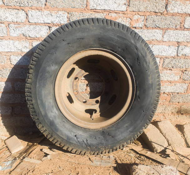 een oude en versleten vrachtwagenband zonder lucht en bedekt met stof en roest rust buiten tegen een betonnen muur omringd door puin en vuil - Foto, afbeelding