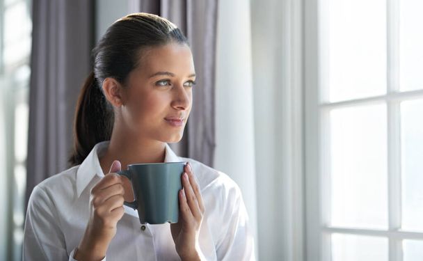 Comienzo del día hábil. Tiro de una joven mujer de negocios disfrutando de una taza de café mientras está de pie junto a un windown. - Foto, imagen