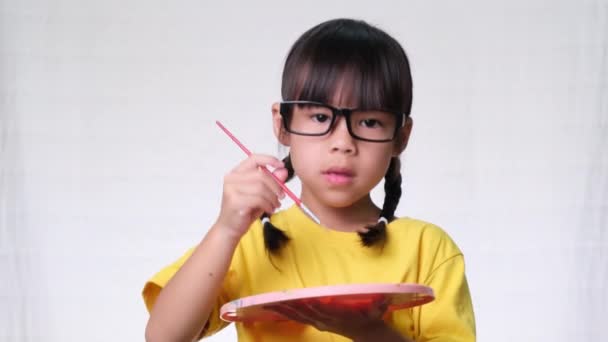 Küçük şirin bir kız elinde fırçayla stüdyoda beyaz arka planda hayali bir resim çiziyor. Eğitim ve hayal gücü kavramı - Video, Çekim