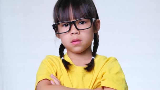 Een boos klein meisje met een bril staat met haar armen gekruist en kijkt naar de camera op een witte studio achtergrond. - Video