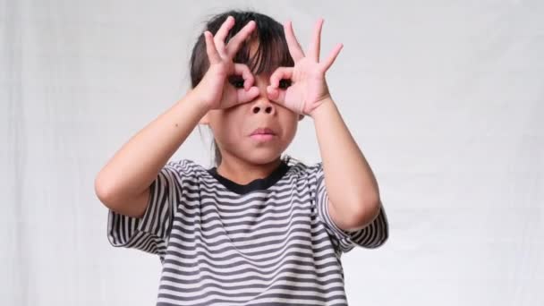 Schattig klein meisje maken oke gebaar met handen op de ogen en kijken door vingers over een witte studio achtergrond. - Video