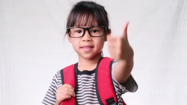 Happy schoolgirl wearing casual outfit with backpack showing thumbs up gesture on white background in studio. Back to school concept - Footage, Video