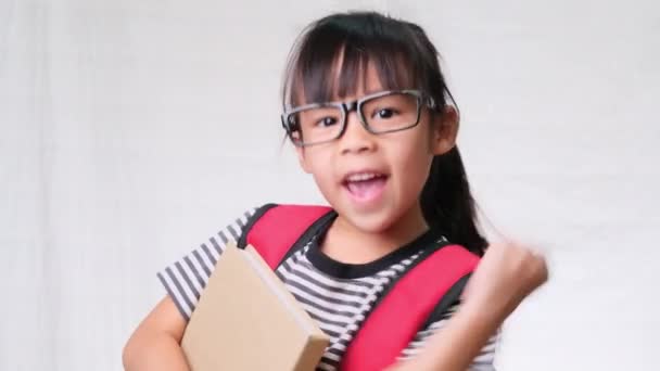 Smiling schoolgirl wearing summer outfit with backpack holding books on white background in studio. Back to school concept - Footage, Video