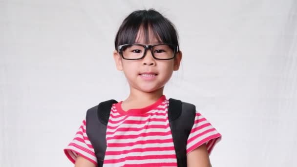 Happy schoolgirl wearing casual clothes with a backpack and doing heart-shaped hands on her head on white background in studio. Back to school concept - Footage, Video