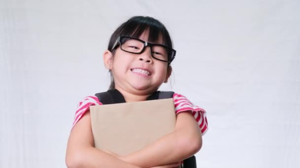 Hymyilevä koulutyttö yllään kesäasu reppu tilalla kirjoja valkoisella taustalla studiossa. Takaisin kouluun käsite - Materiaali, video