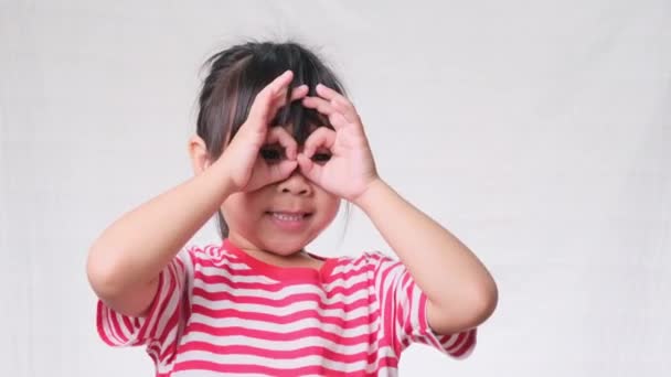 Aranyos kislány vicces arcokat visel ujjazó szemüveg, alakú, mint néz keresztül távcső egy fehér stúdió háttér. - Felvétel, videó