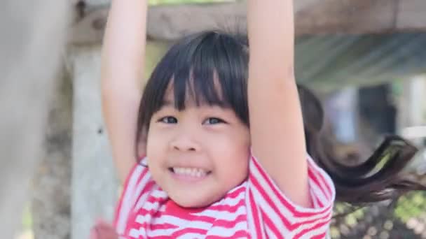 Linda niña asiática cuelga una barra de madera con sus manos para hacer ejercicio en el patio trasero. Niño activo cuelga en la barra con dos manos. - Metraje, vídeo