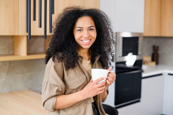 Waist up portrait view of a beautiful multiracial brunette girl standing with a cup of coffee in her hands at the kitchen and smiling - Zdjęcie, obraz