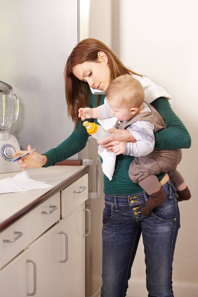 Being a mom is a real juggling act. Young mom trying to multi-task while holding her baby son and his bottle. - 写真・画像